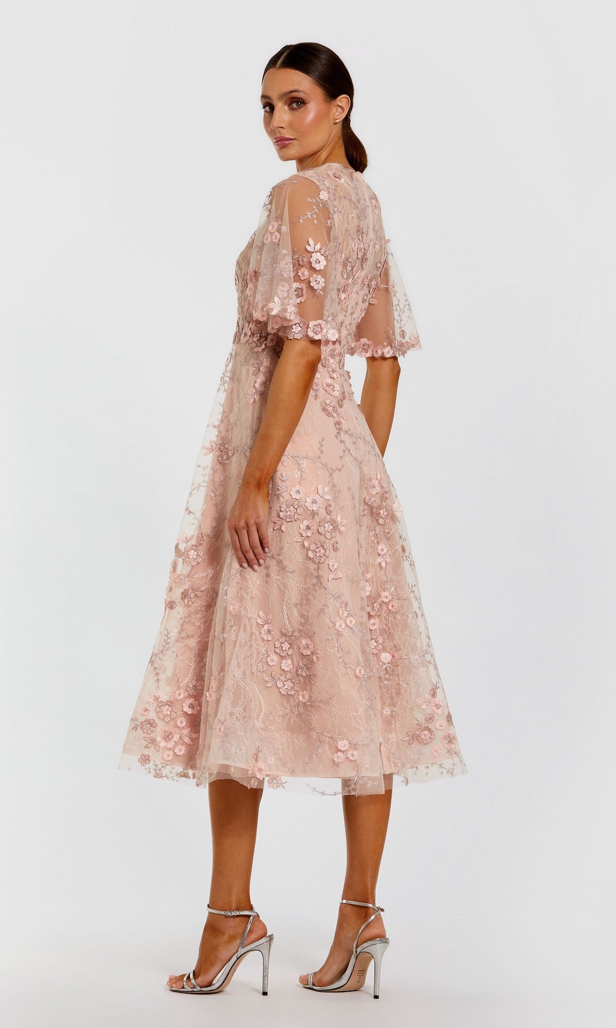 A woman stands against a plain background wearing the Mac Duggal 69036—a blush pink, knee-length, floral-embroidered mesh midi wedding guest dress with sheer sleeves—paired with silver high-heeled sandals.